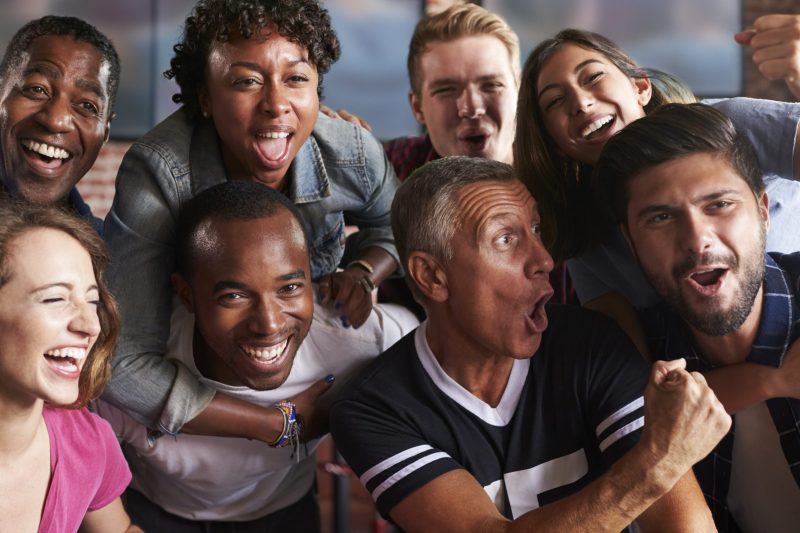 Portrait Of Friends Watching Game In Sports Bar On Screens
